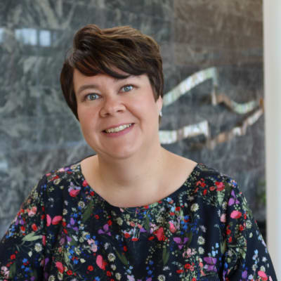 Tummahiuksinen nainen kukikkaassa paidassa hymyilee suoraan kameraan. 