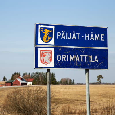 Orimattilan kaupunkikyltti ja Päijät-Hämeen maakuntakyltti päällekkäin maantien varrella. Taustalla paljas, keväinen maalaismiasema.