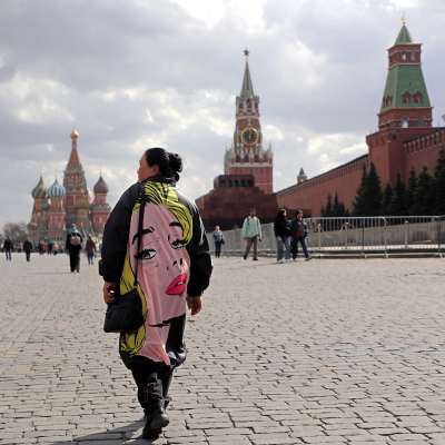 En kvinna går på Röda torget i Moskva med ryggen mot kameran. Till höger syns Kremls röda tegelmur och längre fram Vasilijkatedralens lökkupoler. På torget står människor här och dä och fotograferar. Kvinnan är klädd i en kappa som på ryggsidan har ett tecknat kvinnoansikte med röda läppar och blont hår.