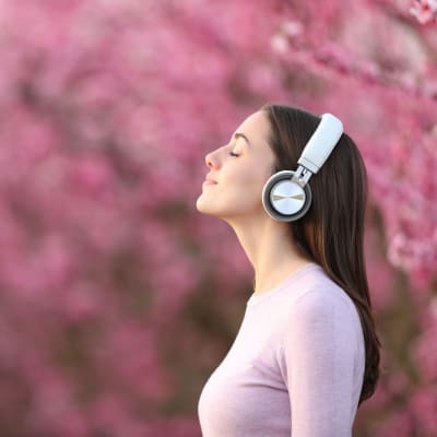 En kvinna med hörlurar på sig fotograferad från sidan. Hon står utomhus i ett fält av rosa blommor och träd.