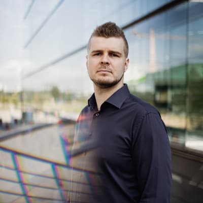 Mies seisoo Tampereen ylopiston ikkunan edessä ja katsoo vakavan kameraan. 