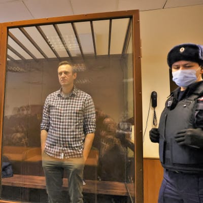 Aleksei Navalnyi seisoo ködet taskuissaan lasiseinäisessä häkissä oikeussalissa.Hänellä on ruudullinen paita. Häkin edessä seisoo univormupukuinen mies.
