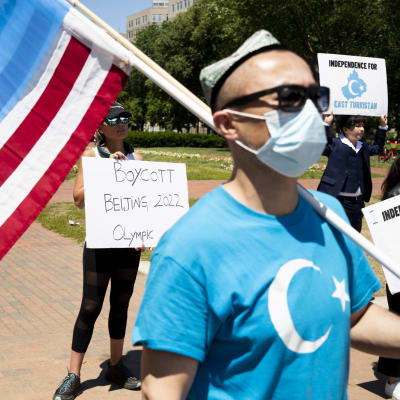 En man i munskydd och solglasögon bär både den amerikanska och uiguriska flaggan över axeln. Bakom honom syns en kvinna med ett plakat med texten "Boycott Beijing 2022 Olympic".
