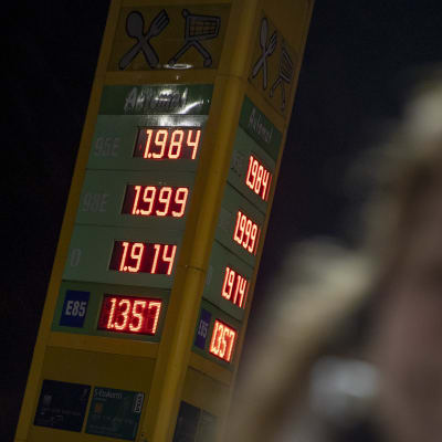 Minna Jaala seisoo huoltoaseman pihalla. Taustalla polttoaineiden hinnat. 