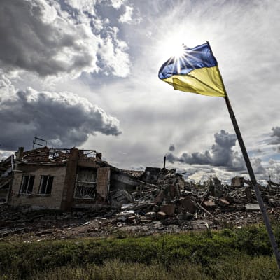 Den ukrainska flaggan vajar i motljus, i bakgrunden ett bostadshus i ruiner. 