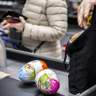 Ihmisiä pakkaamassa kassalla ostoksia. Kaksi pääsiäismunaa vielä pakkaamati.