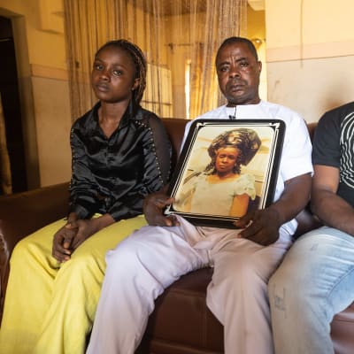 Nigerialaiset aikuiset tytär, isä ja poika istuvat sohvalla, isän sylissä perheen kuolleen äidin valokuva, 
