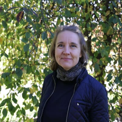Eläinlääkäri Kati Lehtimäki seisoo puun alla, päällään sininen takki ja kaulassa huivi. Hymyilee kuvaajalle. 