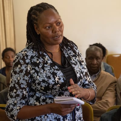 Kenialainen keski-ikäinen nainen puhuu vihko kädessä seisaaltaan kokouksessa, taustalla kenialaisia miehiä istumassa. 