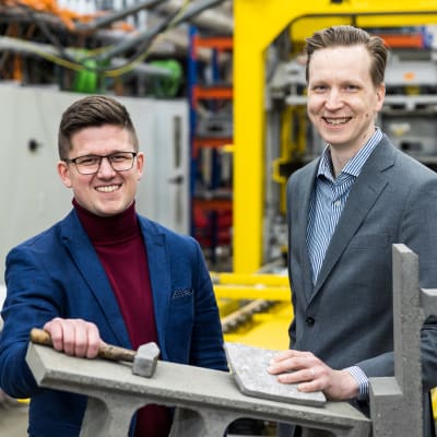 Kaksi pikkutakkeihin pukeutunutta miestä hymyilee betonitehtaalla, betonipalkkien välissä.