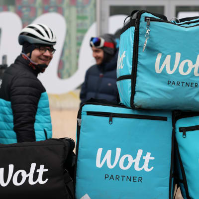 Wolt-lähettien ruokareppuja kasattuna Wolt-Marketin eteen lähettien mielenilmauksessa Kampissa. Taustalla hymyilevä lähetti.