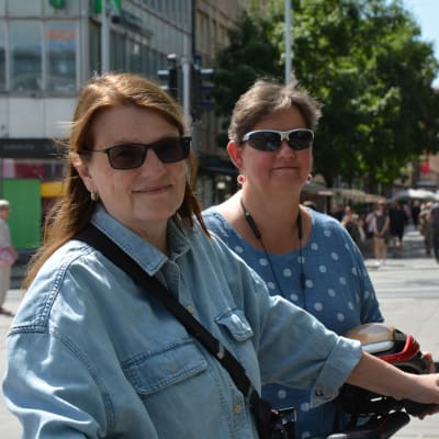 Två kvinnor i blåa tröjor och solglasögonen står med sina cyklar framför en gågata där många människor går