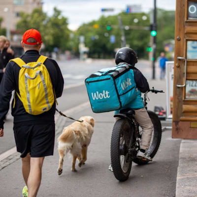 Ruokalähetti seisoo pyöränsä kanssa ravintolan edessä, ohi kävelee mies koirineen.