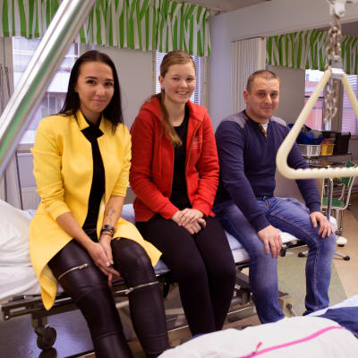 Kseniya Popov, Sara Kinnunen ja Eldar Jandahanov istuvat hoitoluokassa hoitosängyn päällä ja hymyilevät kameralle.