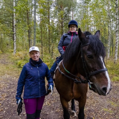 Kaksi naista ja hevonen metsäpolulla. Toinen nainen istuu hevosen selässä.