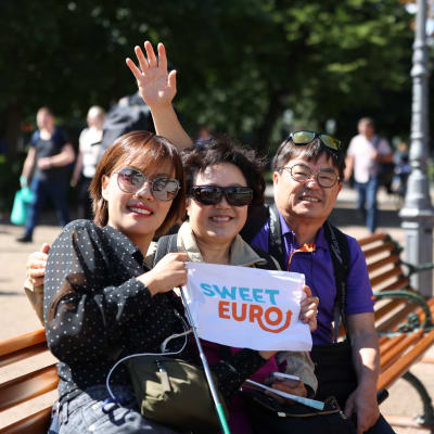 Etelä-Koreasta saapuneet kolme turistia istumassa Helsingin Espan lavan yleisöpenkillä ja katsomassa kameraan. Keskellä heillä on valkoinen lippu, jossa on tekstiä.