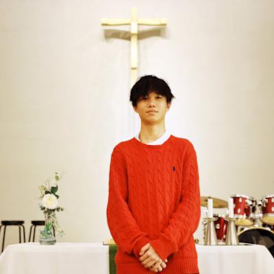 Nuori mies punaisessa neuleessa katsoo kohti. Taustalla risti seinällä.