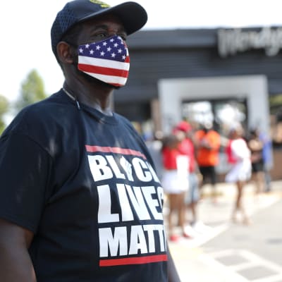 En svart man med ett munskydd i den amerikanska flaggans färger. På mannens skjorta kan man läsa texten Black lives matter.