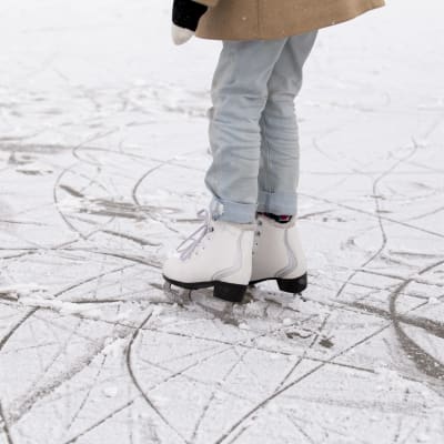 Ett barn med ljusa jeans står på isen i ett par vita skridskor.