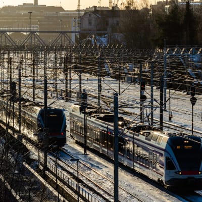 Kaksi junaa kovassa talvisessa valossa ajavat leveää ratapihaa Helsingin päärautatieaseman ja Mall of Triplan välisellä osuudella.