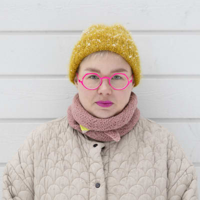 Anni Saukkola seisoo lumisateessa ja katsoo suoraan kameraan. Hänellä on pinkkiä huulipunaa ja silmälasin kehykset. Yllään hänellä on talvitakki, huivi ja pipo. 