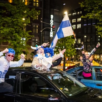 Personer viftande med finska flaggor och iklädda finska färger hänger ut ur fönstret på två bilar i centrala Helsingfors. 