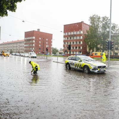 En polis i signalgul regnrock står i knädjupt vatten en bit ifrån en polisbil på en översvämmad gata.