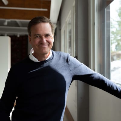 Mika Ihamuotila står vid ett fönster lutandes mot fönsterkarmen. Han ler rakt in i kameran.