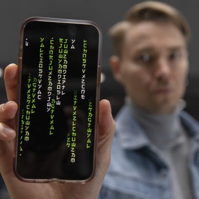 Kuvan etualalla on älypuhelimen näyttö, jossa näkyy graafisia merkkejä. Kännykkää pitelee kädessään epätarkaksi jätetty farkkutakkinen mies.