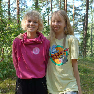 Ungdomarna Ada Willför och Beda Bergsröm står leende i en grön skog. Beda har armen om Adas axel.