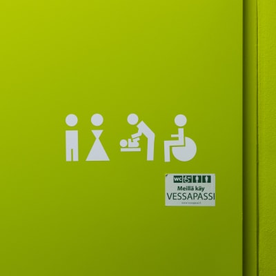 Vihreällä taustalla valkoisia symboleita: Mies, nainen, lastenhoito, pyörätuoli. Niiden alla tarra, jossa lukee "Meillä käy vessapassi".
