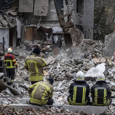 Pelastushenkilöitä istuu tuhoutuneen kerrostalon edessä.