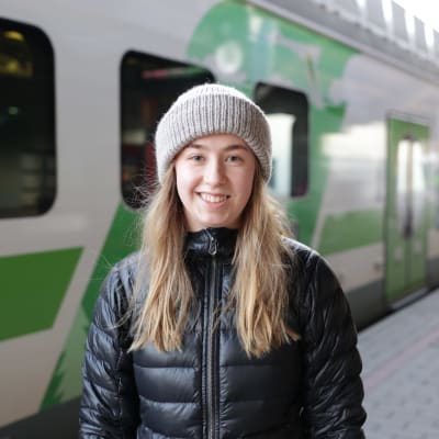 Emma Hannibal seisoo Tampereen rautatieaseman laiturilla junan vieressä, katsoo hymyillen kameraan päällään musta takki ja harmaa pipo.