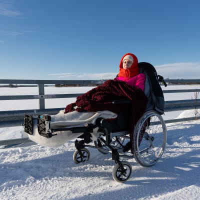 Lilja Ritmos istuu talvella pyörätuolissa talvivaatteissa ja makuupussissa Ketvelniemen ja Kermanniemen välisellä harjulla Kivisalmen sillalla Savitaipaleella.