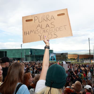 Demonstranter utanför riksdagshuset med skylt som kritiserar Riikka Purra.