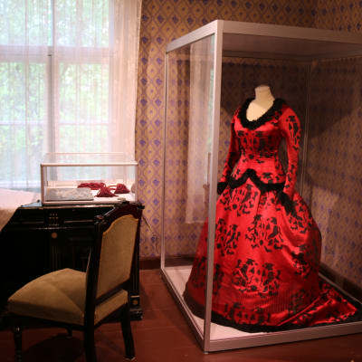 Museossa pöytä, tuoli sekä lasivitriinissä punainen juhlapuku.