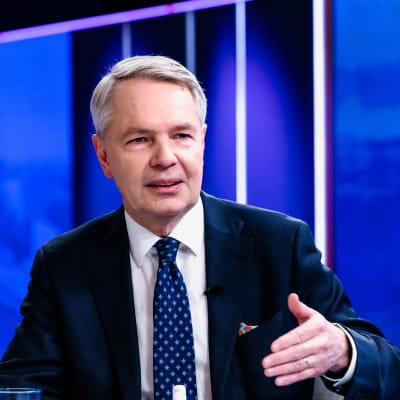 Pekka Haavisto i kostym mot en blå bakgrund viftar med vänster hand.