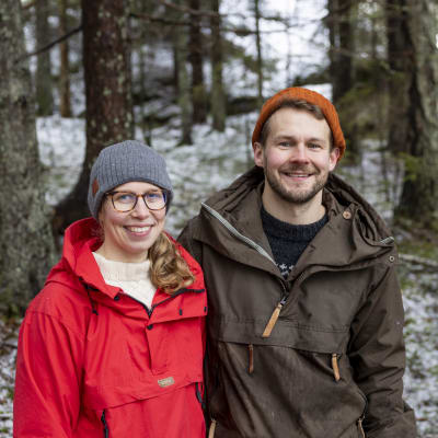 Anna Haavikko ja Joel Heino seisovat vierekkäin ja hymyilevät kameralle. Taustalla ohuen lumikerroksen peittämää kuusimetsää.