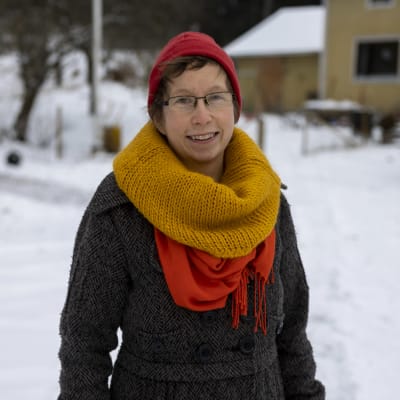 Nainen seisoo talon pihalla ja katsoo hymyillen kameraan.