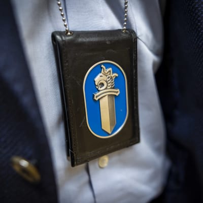 En polisbricka, ett tjänstemärke med lejonet och ett svärd, hänger i en kedja mot en ljusblå polisskjorta.