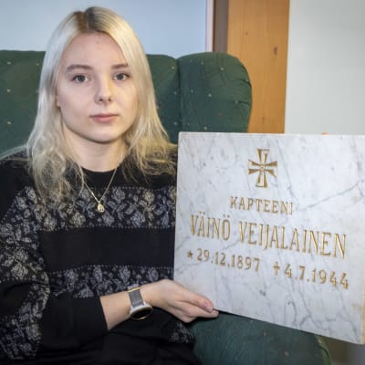 Kaisa Oksa istuu nojatuolissa Väinö Veijalaisen hautakiven kanssa.