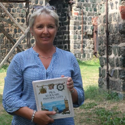 Carina Wolff-Brandt står bland gamla slaggstensbyggnader med sin nya bok i händerna och ser glad ut.