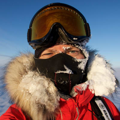 En kvinna iklädd en tjock vinterjacka med pälskantad huva, en balaklava som täcker allt utom ögonen och stora glasögon som hon för tillfället dragit upp på huvudet. Hon befinner sig på Grönland. Huvan är täckt av snö.