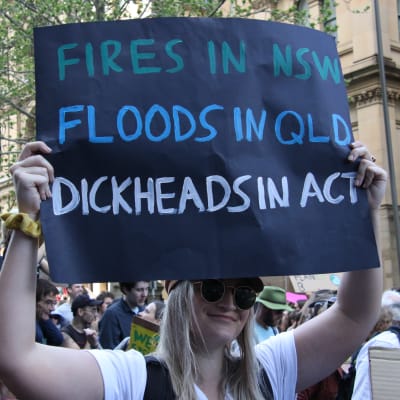 En klimataktivist håller upp ett plakat med texten "Fires i NSW, floods in QLD, dickheads in act".