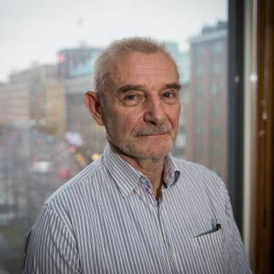 Ralf Sund, en äldre man med kort grått hår och kort grått skägg. Han har en randig skjorta på sig. Han står framför ett fönster med stadsutsikt.