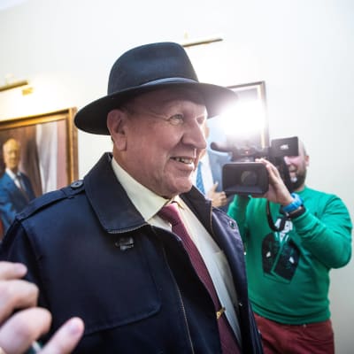 En äldre man i hatt blir fotograferad och har mikrofoner framför sig.