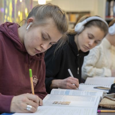 Två studerande antecknar i häften med hörlurar på öronen i biblioteket i Svenska social- och kommunalhögskolan. 