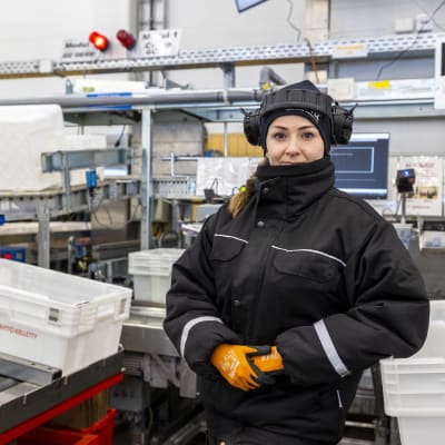 Janina Peräsalo katsoo kameraan työpisteensä edessä. Takana näkyy linjasto, jossa on pakkauslaatikoita.