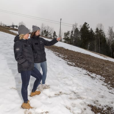 Två personer ser på en halvsnöig backe. En av kvinnorna pekar neråt mot någonting som inte ryms med på bilden.