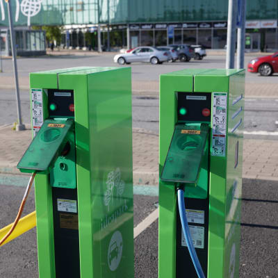 Kaksi sähköautojen latauspistettä. Taustalla aurinkoinen kauppakeskuksen parkkialue, jossa autoja.